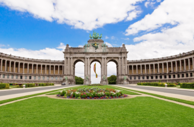 a large stone building with arches and a circular lawn with Cinquantenaire in the background